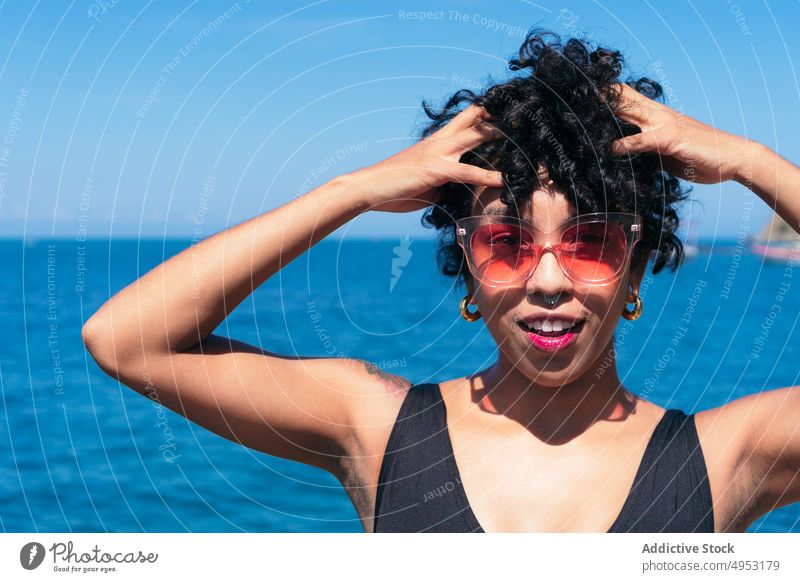 Lächelnde schwarze Frau mit Sonnenbrille vor endlosem Meer heiter Individualität Stil Tattoo feminin Hände auf dem Kopf MEER Blauer Himmel modern kreativ Design