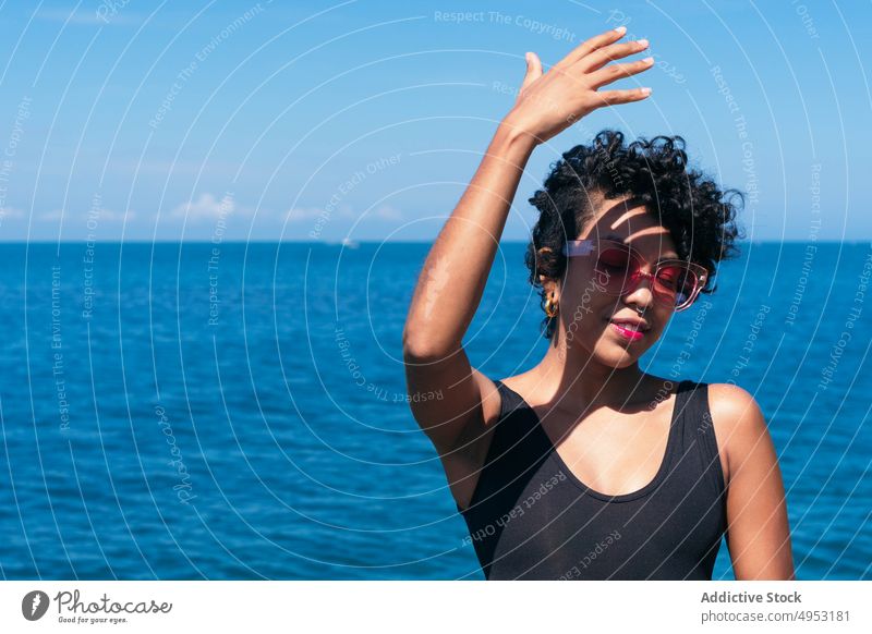 Lächelnde schwarze Frau mit Sonnenbrille vor endlosem Meer heiter Individualität Stil Tattoo feminin Schatten MEER Blauer Himmel modern kreativ Design