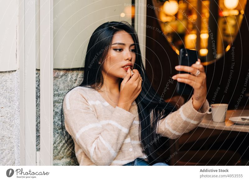 Asiatische Frau mit Smartphone schminkt sich im Café die Lippen Lippenstift Make-up Gesicht Kosmetik Schönheit feminin Fenstersims benutzend Apparatur Gerät