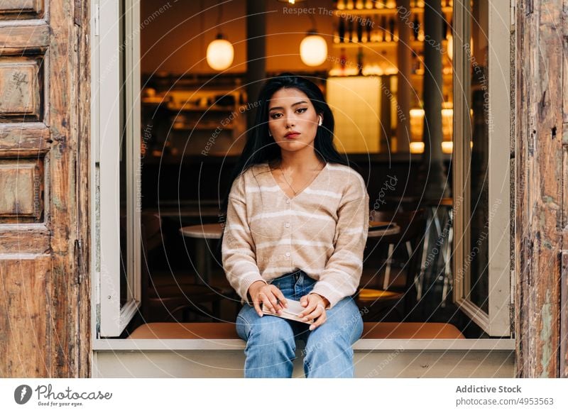 Asiatische Frau mit Ohrstöpseln, die sich auf der Fensterbank eines Cafés ausruht feminin Stil lässig romantisch verträumt Fenstersims hölzern Material alt