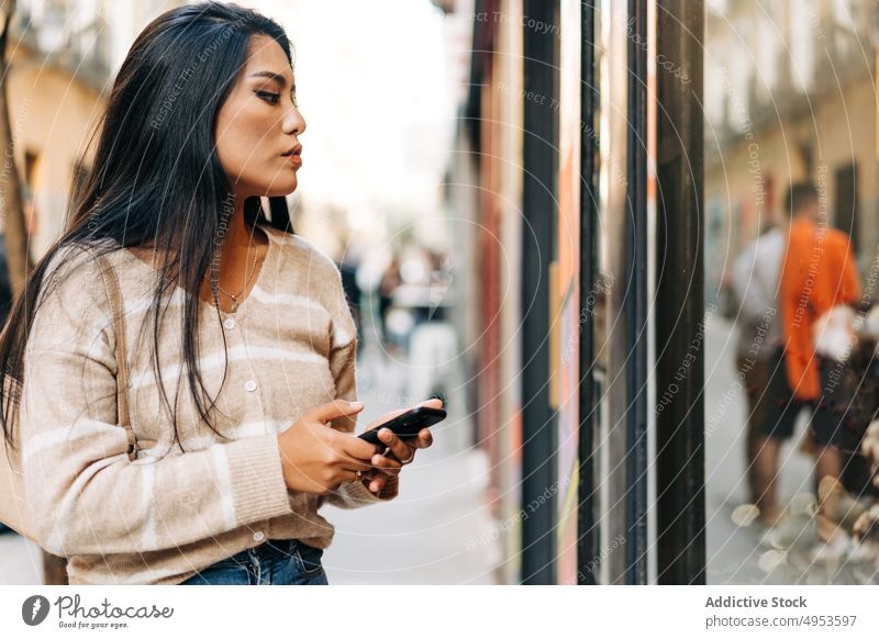 Asiatische Frau mit Smartphone vor einer Glaswand in der Stadt Reflexion & Spiegelung nachdenklich feminin Stil lässig Straßenbelag Großstadt achtsam