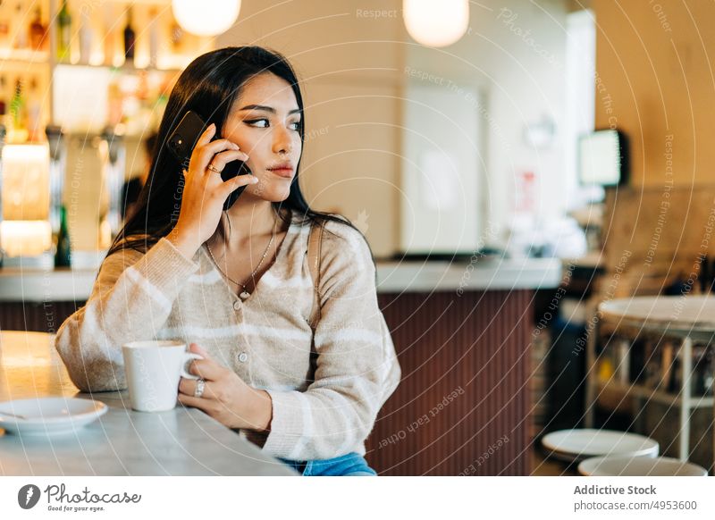 Asiatische Frau mit Kaffee und Smartphone am Tisch eines Cafés sprechend Internet online Freizeit benutzend Apparatur Gerät Funktelefon Tasse Untertasse