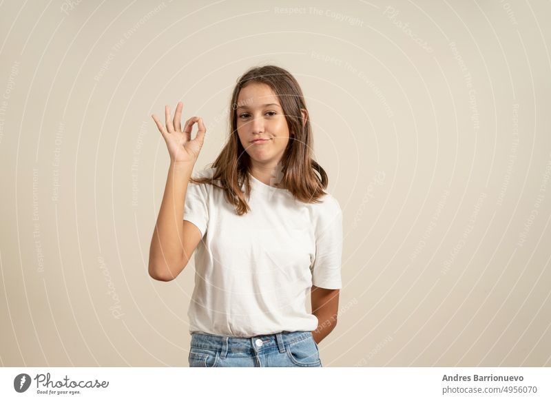 Porträt eines fröhlichen brünetten Teenagers in weißem T-Shirt, der ok singt, isoliert auf beigem Hintergrund. OK hübsch vereinzelt Mädchen jung Frau niedlich