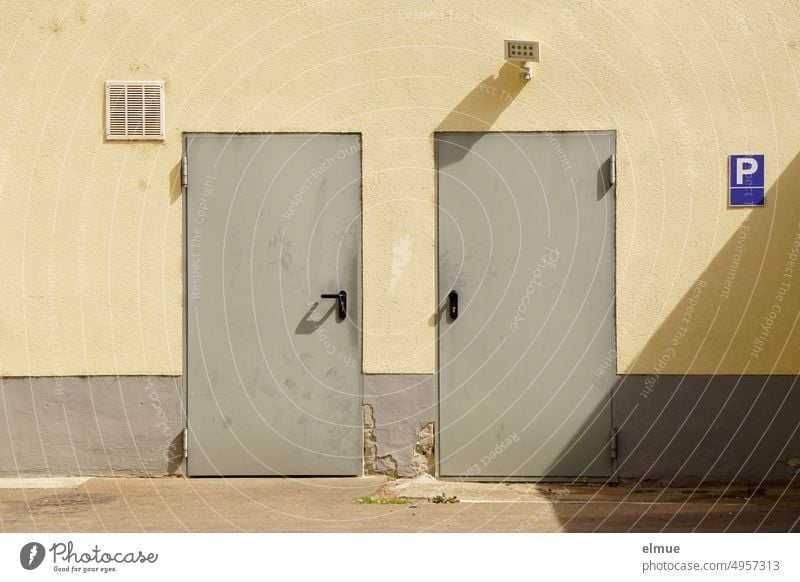 zwei graue, abgegriffene Metalltüren führen in ein Zweckgebäude an dessen Wand sich ein Lüfter, ein kleiner Scheinwerfer und ein blaues Parkplatzschild befinden