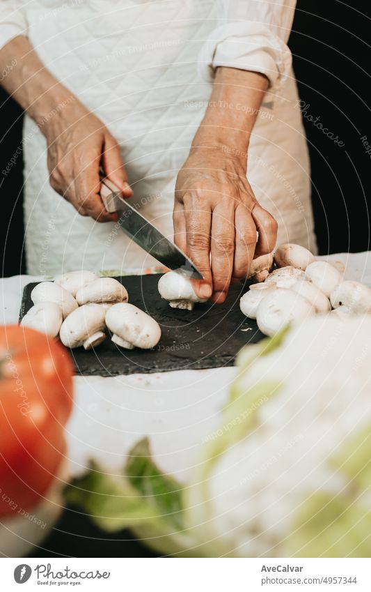 Die faltigen Hände einer älteren Frau, die mit hochgekrempelten Ärmeln auf einem schwarzen Brett Pilze schneidet und die richtige Art des Schneidens lehrt, um ein vegetarisches Gericht mit Tomaten und Blumenkohl zu kochen