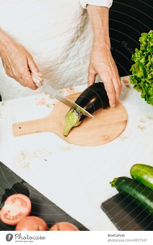 Ältere Köchin beim Kochen eines veganen Rezepts für eine Fernsehsendung. Hände halten ein Messer und schneiden eine Aubergine zusammen mit Zucchini, Tomaten und Salat auf einem Holztisch - eine ausgewogene und gesunde Mahlzeit