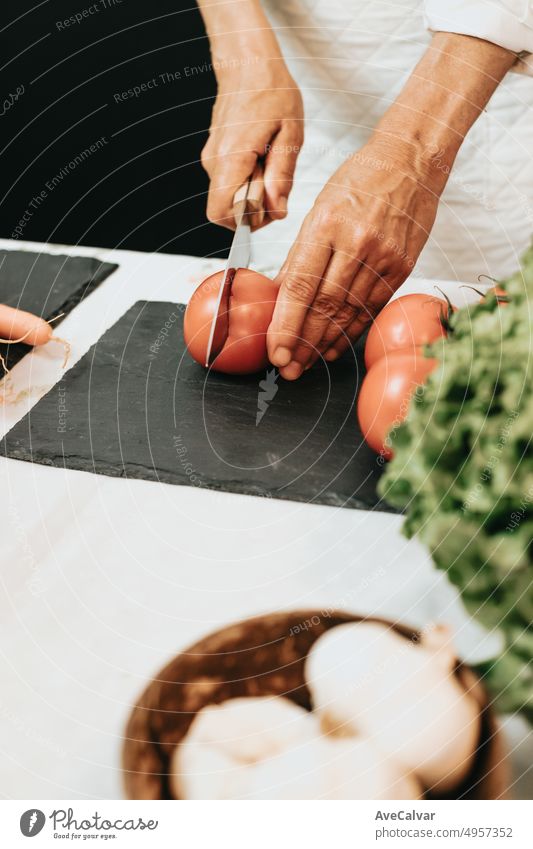 Alte Frau unterrichtet ein Kochrezept mit einer weißen Küchenschürze. Nahaufnahme von Händen schneiden Tomaten mit einem Messer auf einem schwarzen Brett der Vorbereitung einer gesunden, leckeren und komplette Mahlzeit für Mittag-und Abendessen