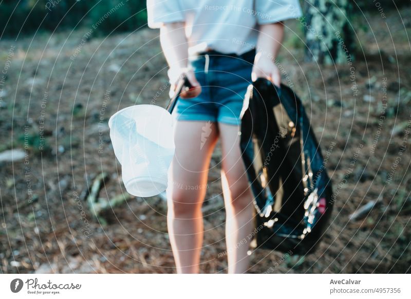 Junge Frau beim Aufsammeln von Müll mit einer Müllzange, die auf die Kamera zeigt.Waldhintergrund.Plastikbecher und Müllsack.Neue Generation.Recyceln, Schutz des Planeten, Umweltschutz, Freiwillige