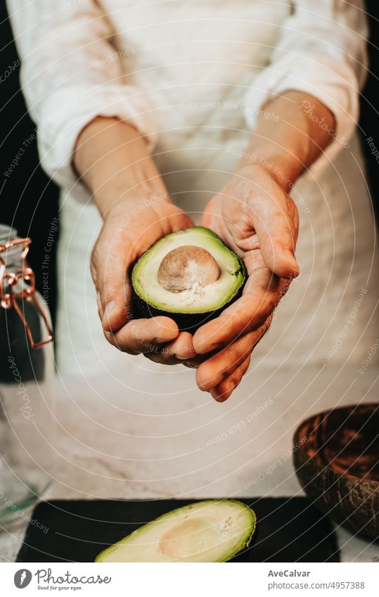 Frau Koch hält und zeigt reife geschnittene Avocado auf weiße Küche apron.Half Avocado auf schwarzem Brett, hölzerne Schüssel für frische Guacamole und Glas für gesunde und nahrhafte Avocado Smoothie