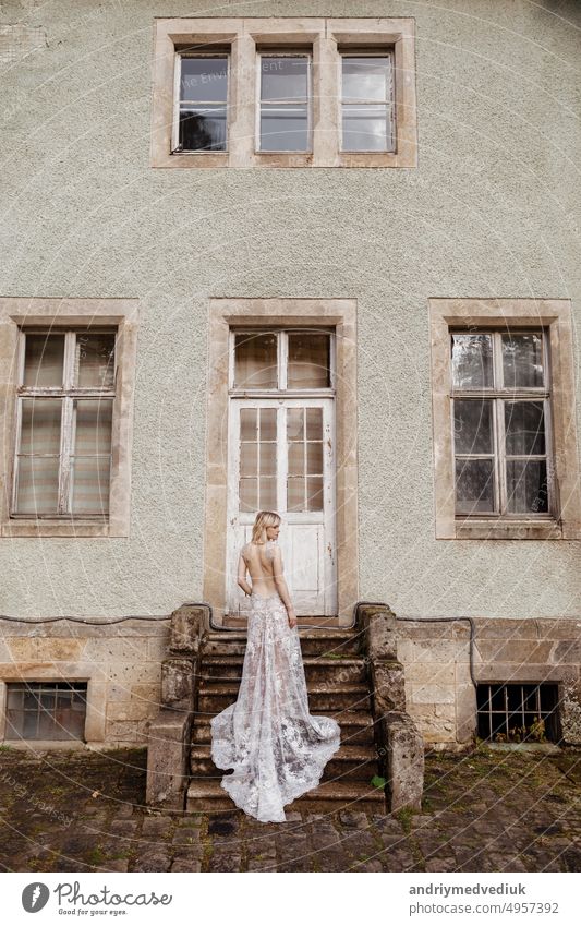 Mode Outdoor-Foto von wunderschönen jungen Braut mit blonden Haaren in eleganten langen Hochzeitskleid posiert in der Nähe der alten Burg. Rückenansicht Kleid