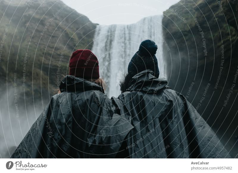 Frauenpaar in Regenkleidung vor dem Skógafoss-Wasserfall in Island an einem stimmungsvollen Tag mit viel Wasser. Lebe deinen Traum, Liebe in Island, Roadtrip-Stil. Besuchen Sie Island.copy space image