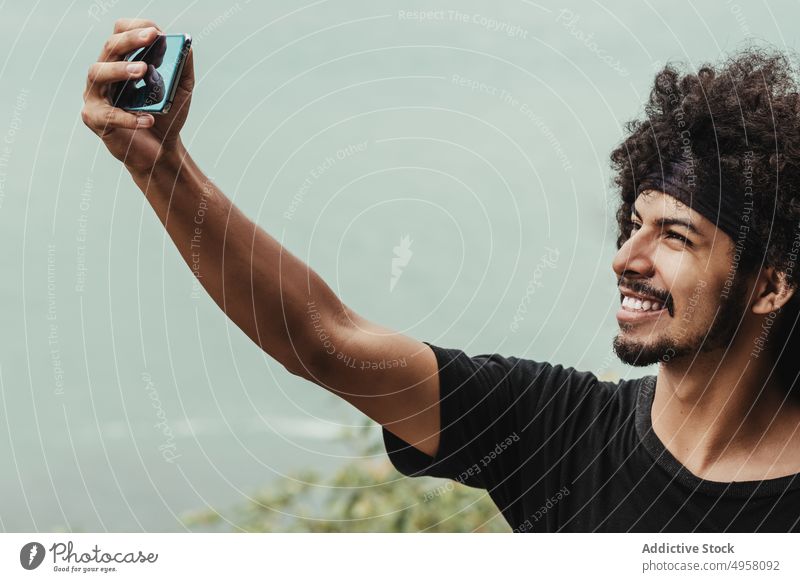Fröhlicher schwarzer Mann, der mit seinem Smartphone ein Selfie gegen das Meer macht heiter Gedächtnis Moment Großstadt Afro-Look benutzend Apparatur Gerät