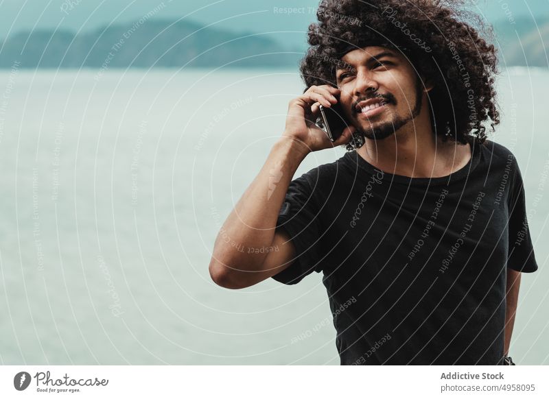 Schwarzer Mann telefoniert mit Smartphone gegen das Meer in der Stadt Telefonanruf Internet online Zeitvertreib interessiert Afro-Look benutzend Apparatur Gerät