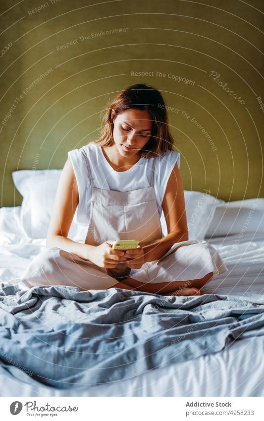 Junge Frau, die auf dem Bett sitzend eine SMS auf ihrem Smartphone schreibt benutzend Textnachricht Nachricht soziale Netzwerke Funktelefon Anschluss Surfen