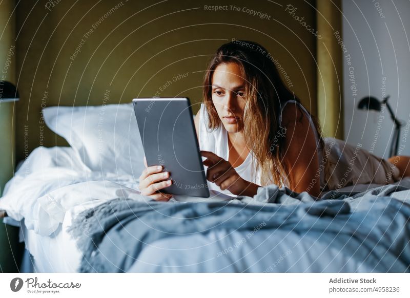 Konzentrierte junge Frau surft auf einem Tablet, während sie auf dem Bett im Zimmer liegt Tablette benutzend Lügen Gerät heimwärts online soziale Netzwerke