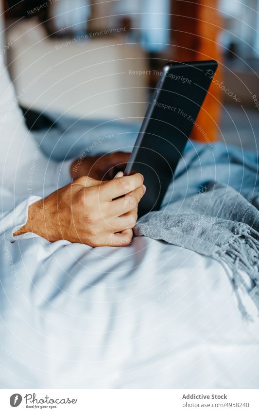 Anonymer Mann surft auf einem Tablet, während er in einem Zimmer auf dem Bett liegt Tablette benutzend Lügen Gerät heimwärts online soziale Netzwerke Surfen