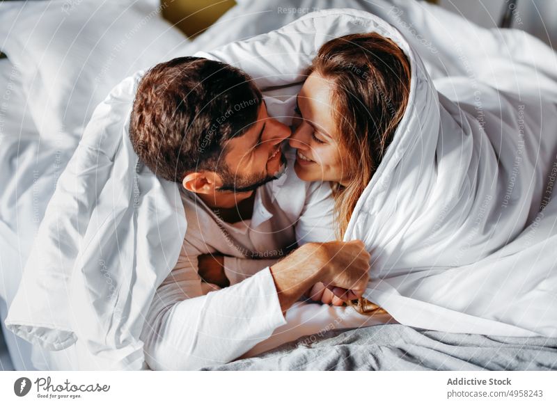 Fröhliches junges Paar, das sich unter einer weißen Decke im Bett streichelt Umarmen Angebot kuscheln Zuneigung Bettdecke Zusammensein Partnerschaft Glück Liebe