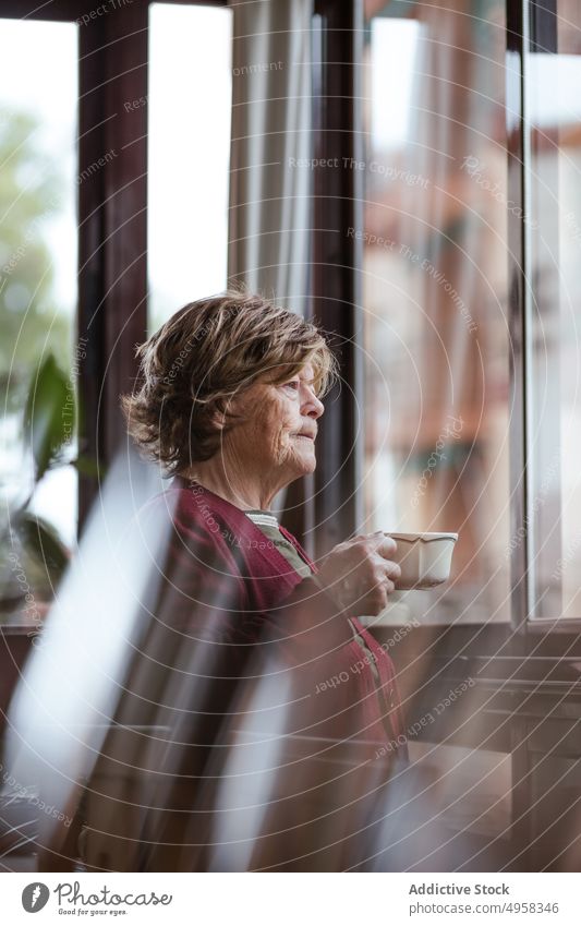 Nachdenkliche ältere Frau trinkt heißes Getränk am Fenster gemütlich trinken Tasse heimwärts besinnlich ruhen in den Ruhestand gehen lässig Raum Lifestyle