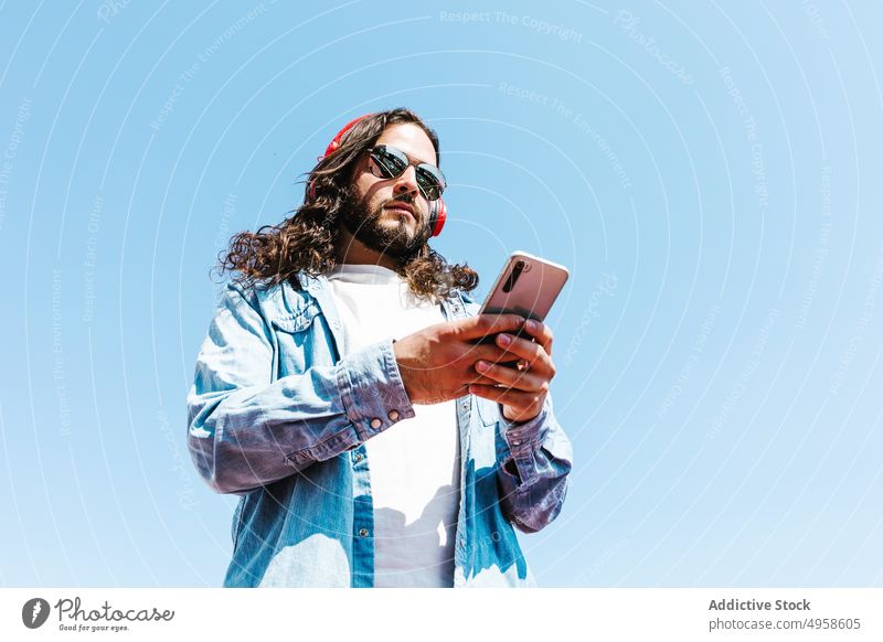 Ethnischer Mann mit Sonnenbrille und Smartphone im Sonnenlicht männlich Stil Browsen Individualität Glück Kopfhörer Lifestyle bärtig benutzend Apparatur Gerät