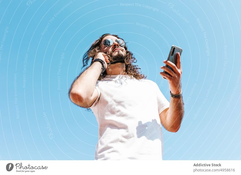 Ethnischer Hipster-Mann mit Sonnenbrille und Smartphone im Sonnenlicht Stil Individualität brutal Lifestyle benutzend Apparatur Gerät Zeitgenosse kreativ Design