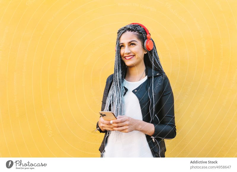 Zufriedene ethnische Frau mit Smartphone, die ein Lied über Kopfhörer hört Musik zuhören Inhalt trendy Geflecht