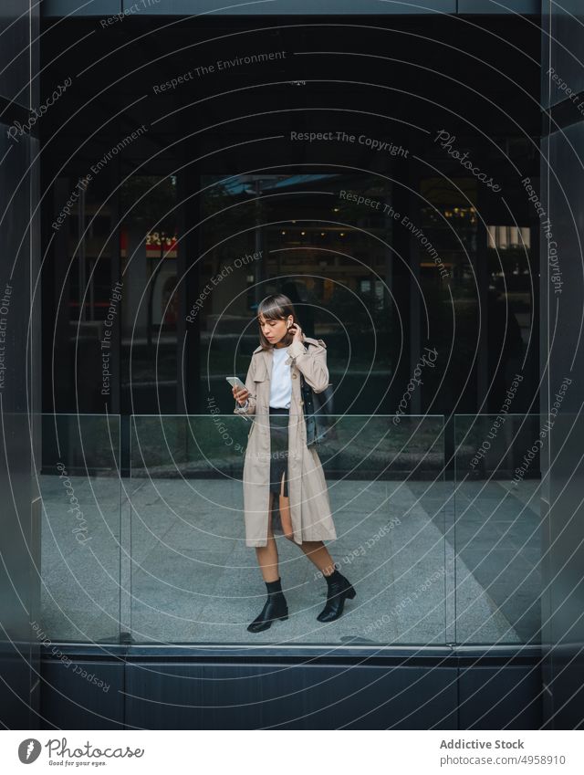 Frau mit Smartphone in der Nähe eines modernen Gebäudes Browsen online Kopfhörer tws Ohrstöpsel urban Straße trendy Outfit Mantel Stil Mode Struktur Lifestyle