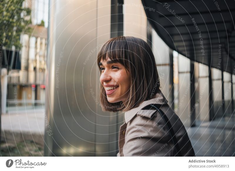 Fröhliche Frau steht in der Nähe eines modernen Gebäudes Stil Terrasse Spalte trendy urban Straße Outfit Mantel Mode Struktur anlehnen Vorschein heiter
