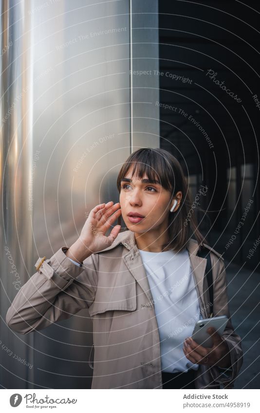 Junge Frau mit Kopfhörern schaut in der Nähe eines Gebäudes weg modern Smartphone sprechend Kommunizieren Gespräch urban Videoanruf trendy Straße Terrasse