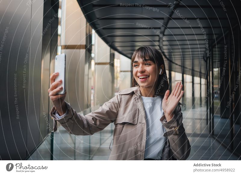 Fröhliche Frau mit Videoanruf in der Nähe eines modernen Gebäudes Kopfhörer Smartphone Video-Chat Kommunizieren Gespräch Wellenhand Straße Gruß urban trendy