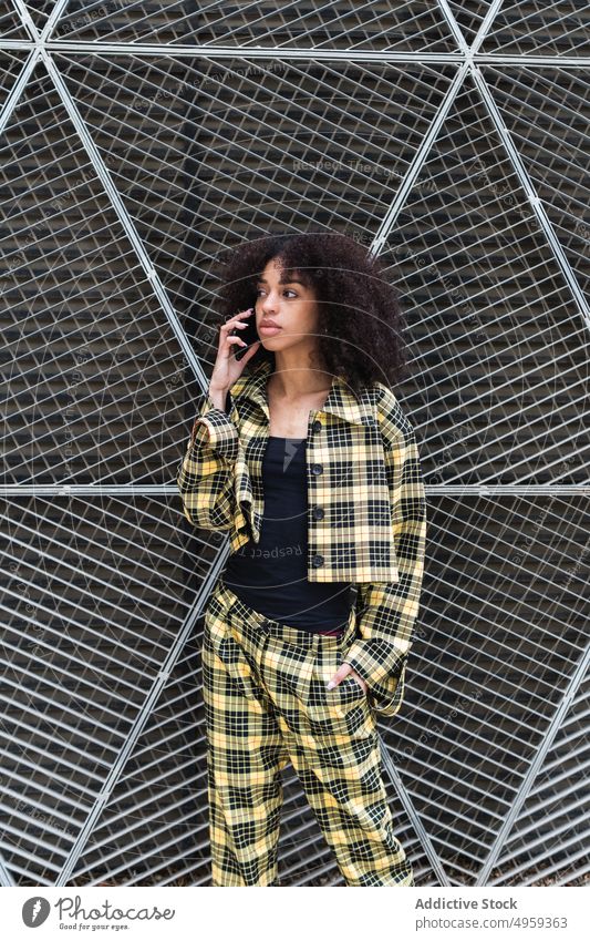 Trendige ethnische Frau, die mit ihrem Smartphone vor einem Gitterzaun spricht reden Stil Mode Hand-in-Tasche Individualität cool feminin benutzend Apparatur