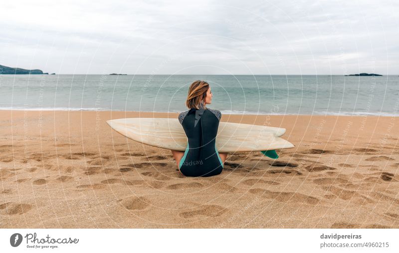 Surferin mit Neoprenanzug und Surfbrett, die auf dem Sand sitzt und zur Seite auf den Strand schaut Frau jung zur Seite schauend Sitzen MEER Winter Rücken