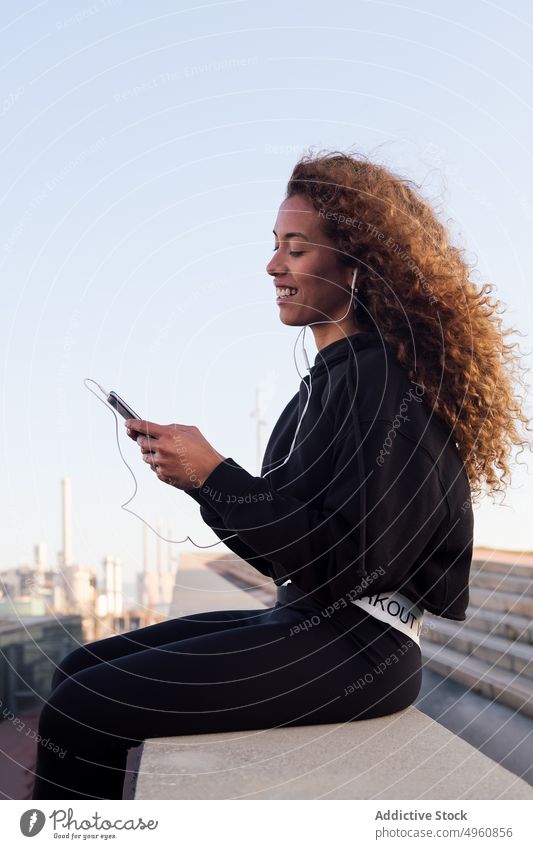 Lächelnde schwarze Sportlerin, die in der Stadt mit ihrem Smartphone chattet plaudernd Kopfhörer Internet online Pause Großstadt benutzend Apparatur Frau Gerät