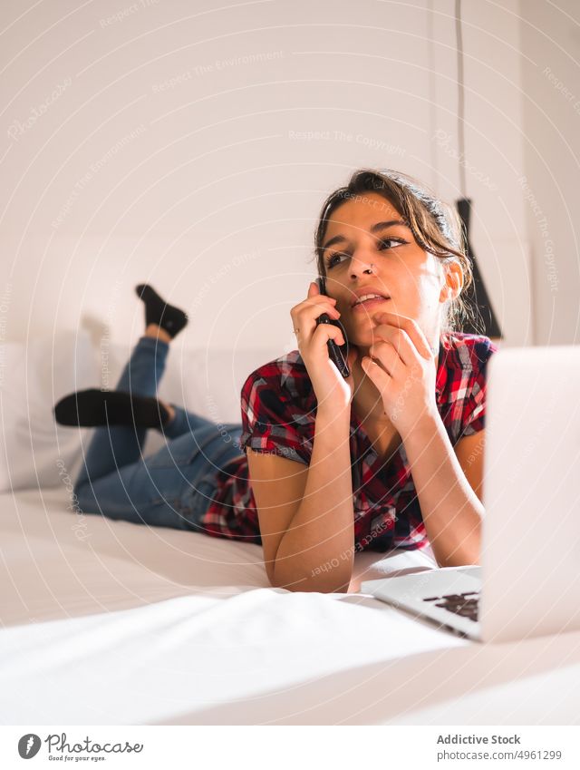 Nachdenkliche Frau, die auf dem Bett mit Laptop auf ihr Smartphone spricht Gespräch Kinn berühren besinnlich freie Zeit Beine gekreuzt benutzend Apparatur Gerät