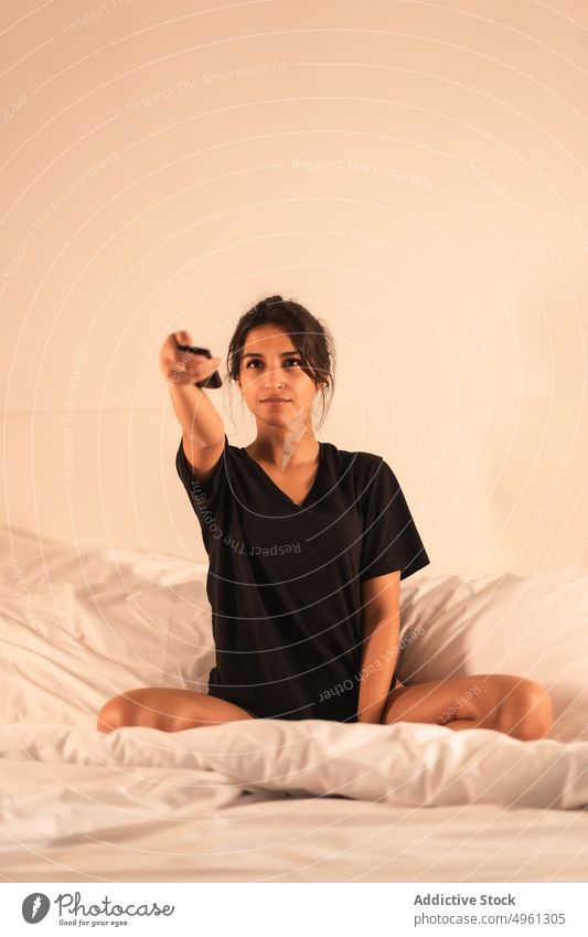 Frau mit Fernbedienung auf ungemachtem Bett selbstbewusst Zeitgenosse Generation genießen ausdehnen Hotel Porträt Gerät Barfuß T-Shirt selbstsicher zerknittert