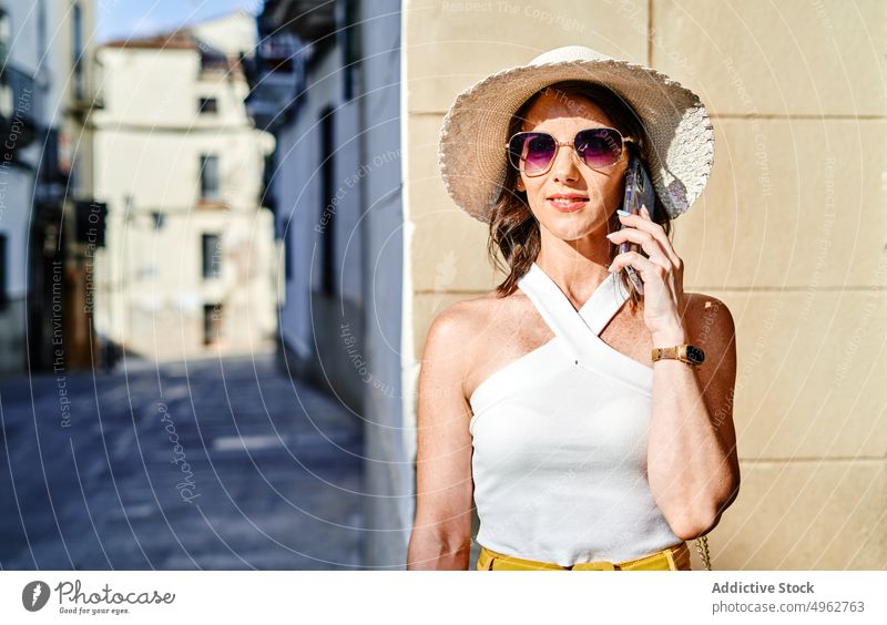 Positive Frau, die in der Stadt mit ihrem Smartphone spricht reden Großstadt Gespräch Telefonanruf Straße Sommer Hut sprechen Mobile Apparatur Sonnenbrille