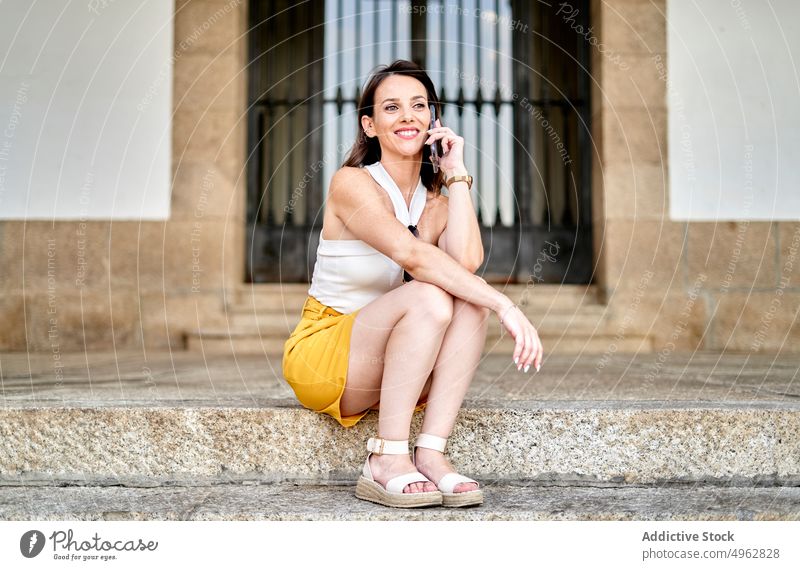 Glückliche junge Frau mit Telefonanruf sitzt auf Schritt auf der Straße reden Smartphone Lächeln Gespräch Kommunizieren Optimist Mobile dunkles Haar Stil