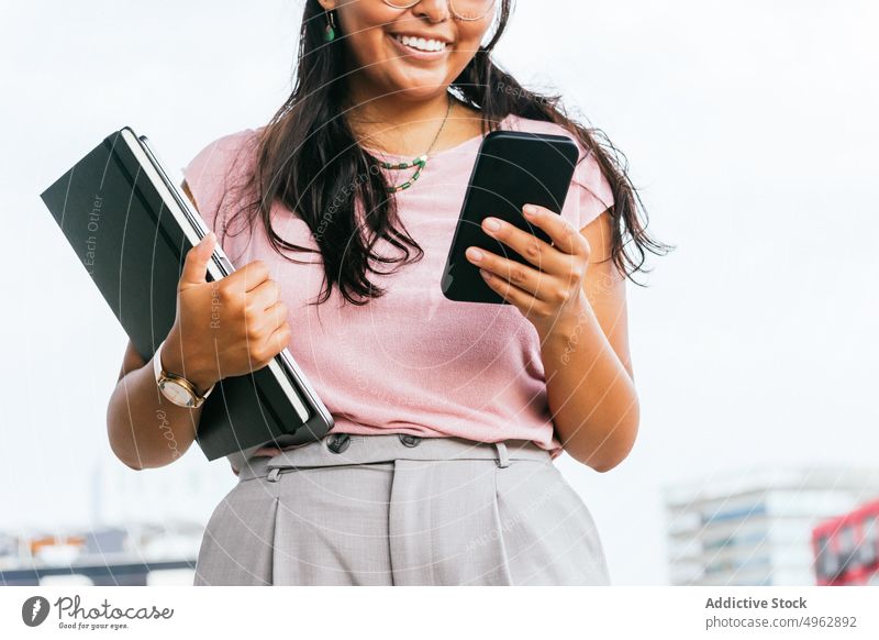 Lächelnde junge ethnische Geschäftsfrau mit Smartphone in der Nähe moderner Gebäude Frau Nachricht Unternehmer positiv Kommunizieren benutzend Internet Straße