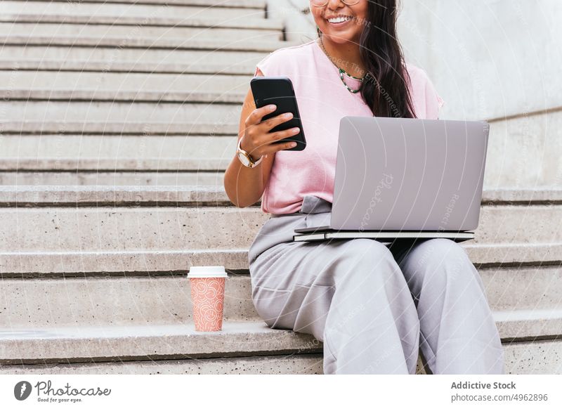 Crop ethnische Frau sitzt auf einer Treppe und benutzt Laptop und Smartphone auf der Straße freiberuflich benutzend Lächeln abgelegen Imbissbude Kaffee Arbeit