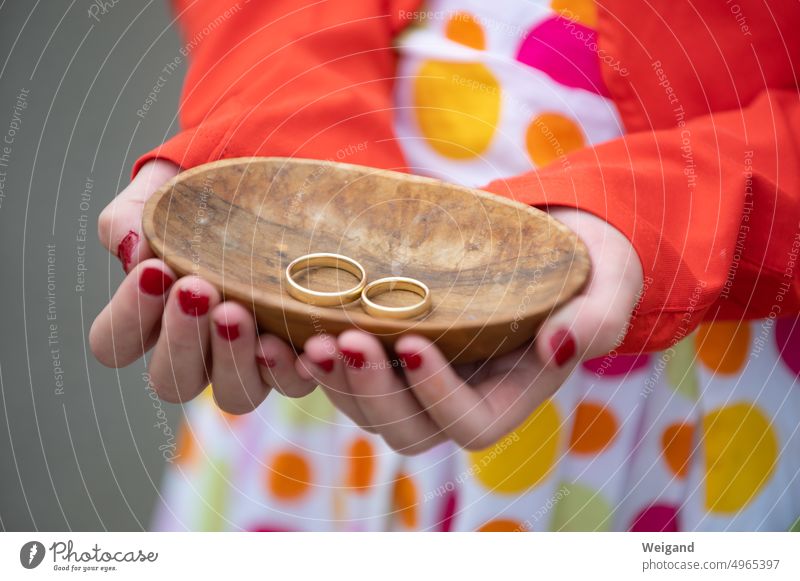 Eine Frau in einem bunt gepunkteten Kleid mit rotem Blazer hält eine kleine ovale Holzschale mit zwei goldenen Ringen in beiden Händen in die Kamera Goldringe