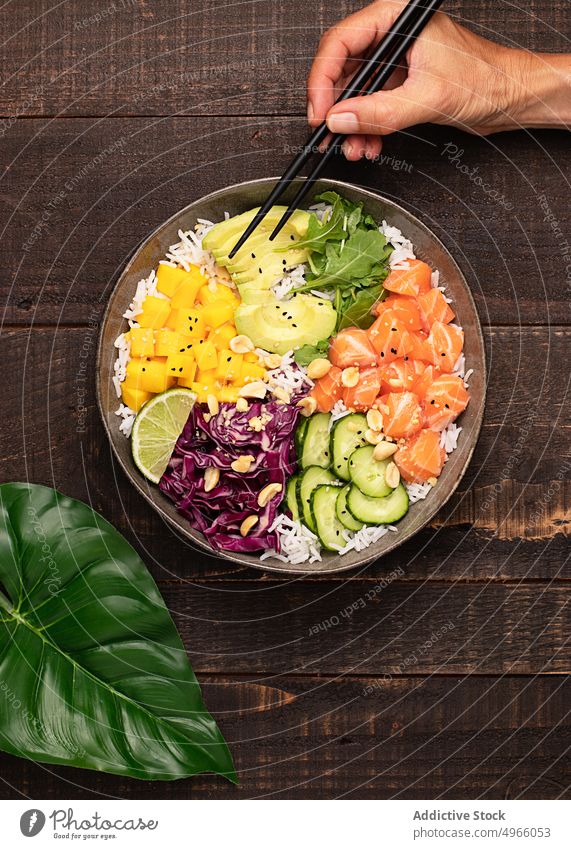 Anonyme Person isst Poke Bowl mit verschiedenen Gemüsesorten, Reis und Lachs stoßen Avocado Mango Asiatische Küche Essstäbchen gesunde Ernährung Salatgurke