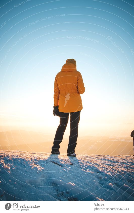 Ein Reisender in einer markanten gelben Jacke steht bei Sonnenuntergang auf einem Hügel in den polnischen Beskiden und genießt den Moment. Echte Menschen in winterlicher Eislandschaft