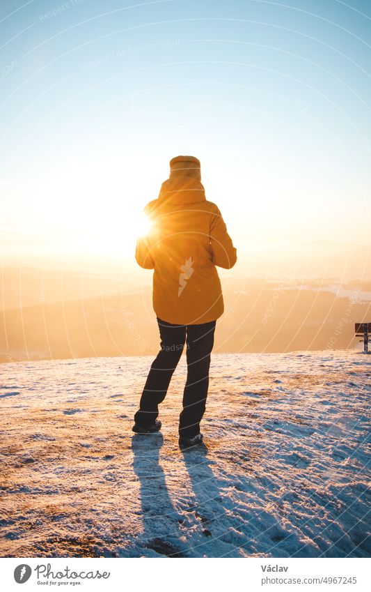 Ein Reisender in einer markanten gelben Jacke steht bei Sonnenuntergang auf einem Hügel in den polnischen Beskiden und genießt den Moment. Echte Menschen in winterlicher Eislandschaft