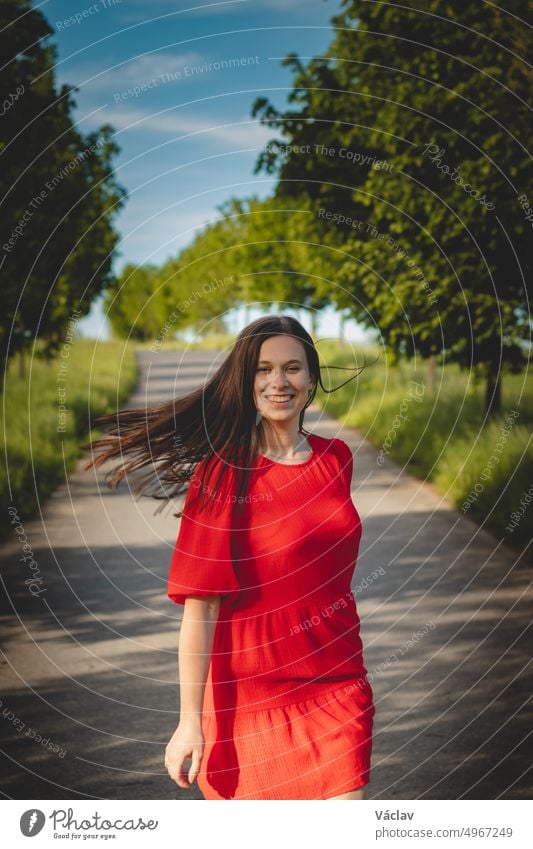 Schöne junge Brünette in einem roten Sommerkleid mit wallendem braunen Haar geht an einem Sommertag auf einem Fahrradweg und in einer Baumallee spazieren. Unverfälschtes Porträt einer Studentin
