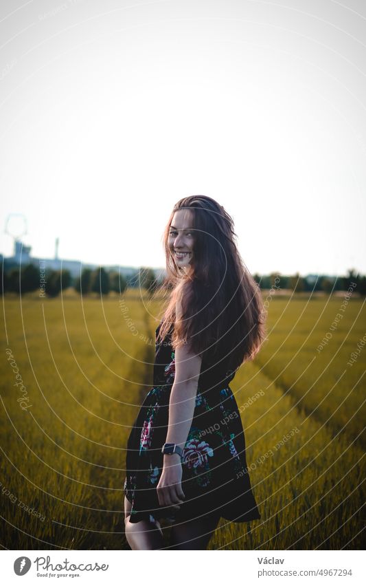 Atemberaubende Brünette Modell mit einem natürlichen breiten Lächeln in einem dunklen Sommer blumigen Kleid Spaziergänge durch das hohe Gras, ihre gewellten dunklen Haare fliegen. Candid Porträt echte erwachsene Frau. Tschechische Republik, Europa