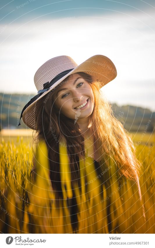 Unverfälschtes Porträt einer jungen Frau aus der Tschechischen Republik, die bei Sonnenuntergang mitten auf einem Feld sitzt und in einem geblümten Kleid die letzten Strahlen genießt. Echtes Lächeln