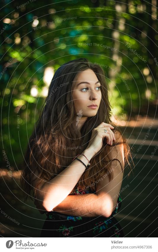 Nachdenklicher Blick einer 24-jährigen Frau mit dunkelbraunem Haar in einem Wald bei Sonnenuntergang. Unverfälschtes Porträt einer hübschen Frau im Sommerkleid
