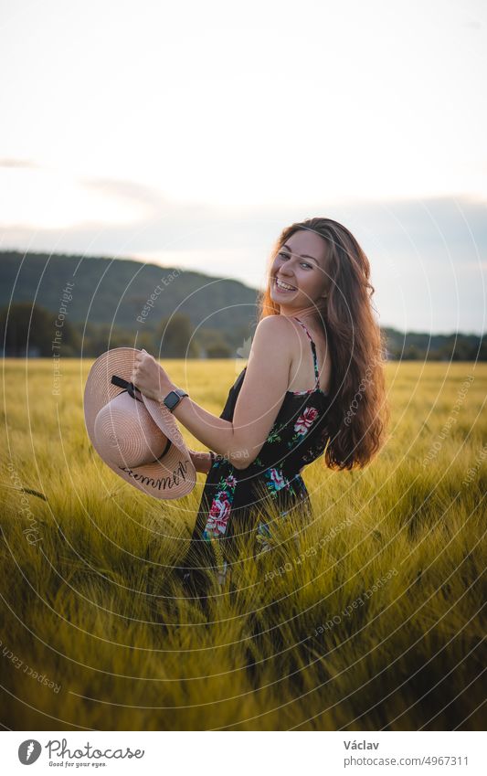 Wunderbares natürliches Lächeln einer erwachsenen Dame in einem geblümten Kleid mit braunem Haar inmitten einer Wiese, die einen schönen Weidenhut trägt. Unverfängliches Porträt eines jungen Modells bei Sonnenuntergang