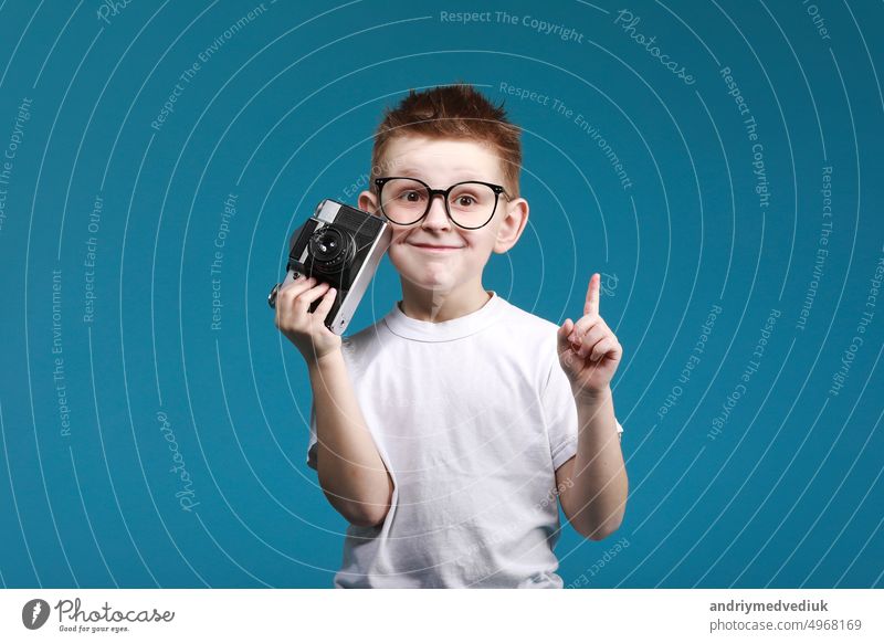 Kleiner Junge, der ein Bild mit einer Retro-Kamera und mit dem Finger nach oben zeigt. Kind Junge mit Vintage-Foto-Kamera isoliert auf blauem Hintergrund. Alte Technologie Konzept. Kind lernt Fotografie