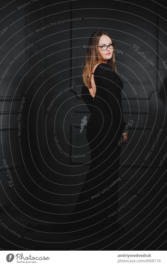 Mädchen in einem langen schwarzen Kleid posiert mit dem Rücken auf einem Hintergrund aus Graphit Wand. Weichzeichner Frau Stehen posierend Schulter offen Seite