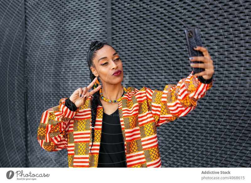 Stilvolle Frau nimmt Selfie auf der Straße Smartphone v-Zeichen schmollende Lippen Wand urban gestikulieren jung ethnisch modern zwei Finger trendy Streetstyle